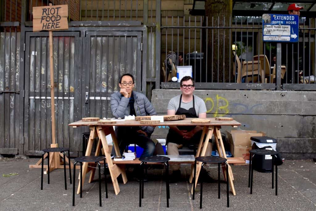 Michael Barnholden and Henry Tsang sit at the table on the street as part of RIOT FOOD HERE