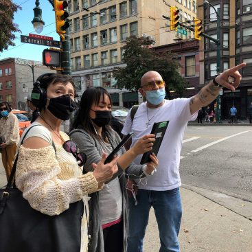 Two women on the left listening to the man on the right who are giving instructions about 360 Riot Walk on Hastings and Carrall Streets